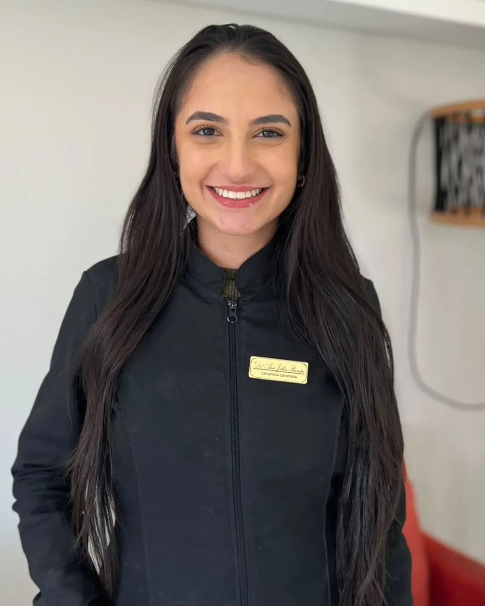 Dra. Ana Júlia Rampe sorrindo no consultório, de jaleco preto com crachá